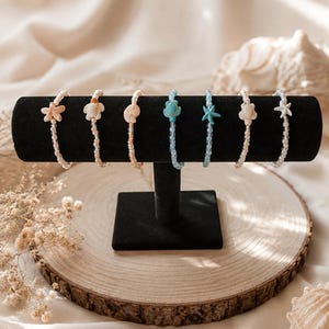 Könnte beinhalten: Sieben Armbänder mit Perlen und Anhängern in Form von Muscheln und Seesternen. Die Armbänder in Weiß, Pfirsich und Blau werden auf einem schwarzen Samtständer auf einer Holzscheibe präsentiert.