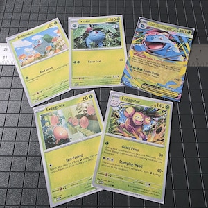 May include: Five Pokémon trading cards, including Bulbasaur, Ivysaur, and Exeggutor, with vibrant illustrations and text describing their abilities and stats. The cards are displayed on a dark, textured surface, likely for a collector or enthusiast.