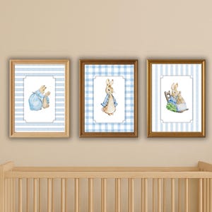 Pode incluir: Três impressões de arte emolduradas para o berçário com ilustrações em aquarela de Peter Rabbit. As impressões têm um esquema de cores azul e branco, com um fundo listrado e xadrez. A obra de arte é exibida acima de um berço de madeira.