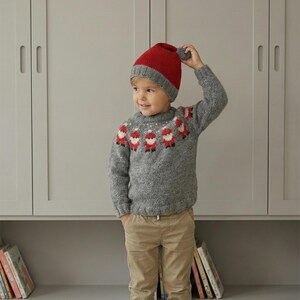 Op de afbeelding: Een kind draagt een grijze gebreide trui met een kerstmanontwerp en een rood-grijze gebreide muts met een pompon. De trui heeft een ronde hals en lange mouwen. Het kind draagt ook een kaki broek.