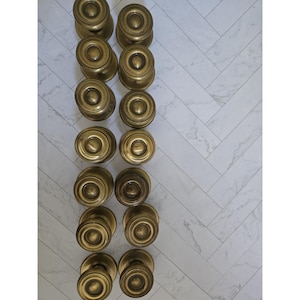 Peut inclure: Une collection de quatorze poignées de porte circulaires en laiton disposées en deux colonnes verticales. Chaque poignée présente une face lisse et arrondie avec un détail circulaire central en relief et une base cylindrique dorée.