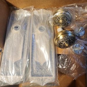 May include: A set of two rectangular door plates and two round door knobs with an antique brass finish and intricate embossed scrollwork patterns. The hardware components are individually wrapped in clear plastic packaging inside a cardboard box.