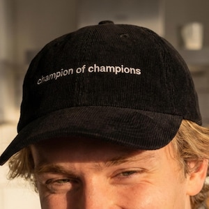 Puede incluir: Gorra de béisbol de pana negra con la inscripción "champion of champions" bordada en blanco. La gorra tiene una visera curva y un diseño clásico de seis paneles. Un accesorio casual para el uso diario.