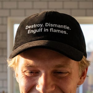 Puede incluir: Gorra de béisbol negra de pana con el texto "Destroy. Dismantle. Engulf in flames." en blanco. La gorra tiene una visera curva y un diseño clásico de seis paneles. La gorra la lleva una persona.