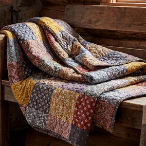 Op de afbeelding: Een veelkleurige patchworkquilt, met tinten geel, rood, blauw en paars, is over een houten bank gedrapeerd. De quilt heeft bloemen- en geometrische patronen. Een mok en een deken liggen ook op de bank, bij een raam.