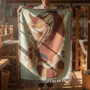 Könnte beinhalten: Eine rechteckige gewebte Wurkdecke mit einem Cartoon-Katzen-Design in Braun-, Beige- und Orangetönen auf hellgrünem Hintergrund. Die Katze hat ein breites Grinsen und steht im Mittelpunkt. Die Decke wird in einer Werkstatt gezeigt.