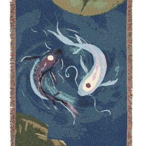 May include: Woven throw blanket featuring a design of two koi fish in a circular pattern against a dark blue background. The fish are in shades of purple, white, and blue. The blanket has a fringed edge with earth-tone colors.