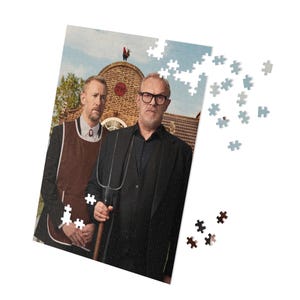 May include: A partially assembled jigsaw puzzle featuring two men in front of a brick building. One man wears glasses and holds a pitchfork. A rooster sits atop the building. Scattered puzzle pieces are visible.