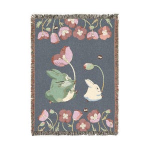 May include: A woven throw blanket featuring a whimsical design with two cartoon rabbits, one green and one white, holding pink flowers. The blanket has a gray background with pink and red flowers and a fringed edge.