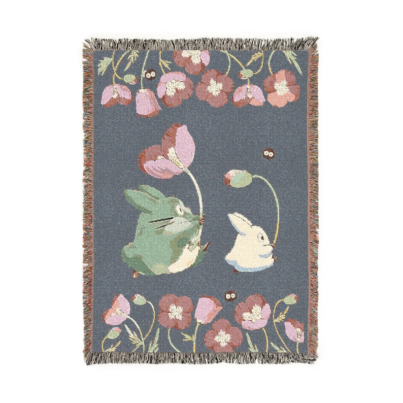 May include: A woven throw blanket featuring a whimsical design with two cartoon rabbits, one green and one white, holding pink flowers. The blanket has a gray background with pink and red flowers and a fringed edge.