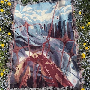 May include: Woven throw blanket featuring an abstract illustration in shades of blue, red, and brown. The design includes stylized figures, clouds, and geometric shapes. The blanket has a fringed edge and is displayed on a bed of flowers.
