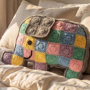 Könnte beinhalten: Handgehäkeltes Elefantenkissen im Patchwork-Design. Das Kissen zeigt Quadrate in Pastellfarben wie Rosa, Blau, Gelb und Grün, mit einem grauen Rand und Ohren. Der Elefant hat ein schwarzes Auge und einen lächelnden Mund.