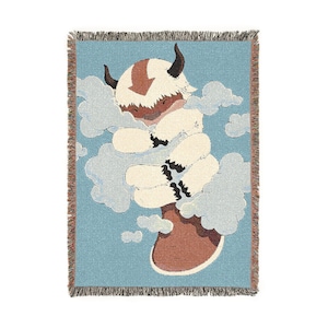 May include: A woven throw blanket featuring a cartoon illustration of a flying bison character in shades of brown, white, and blue. The bison is surrounded by clouds on a light blue background. The blanket has a fringed edge.