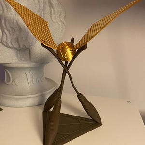 May include: A golden Snitch replica, a fictional object from the Harry Potter series, with detailed wings and a spherical body. The Snitch is mounted on a brown triangular base with two crossed broomsticks.