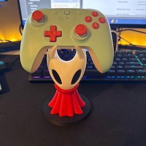 May include: A mint green games controller sits atop a white and red figurine. The controller features red buttons and a directional pad. The figurine has a white head and a red cape, on a black base. The items are on a black surface.