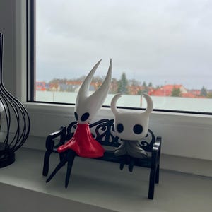 Pode incluir: Duas figuras de personagens do videogame Hollow Knight. Uma figura está vestindo um vestido vermelho e a outra uma capa cinza. Elas estão sentadas em um banco preto em frente a uma janela.
