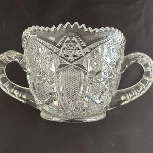 May include: Clear glass sugar bowl with two handles. The bowl features a cut glass design with geometric patterns and a sawtooth rim. The handles are rounded and attached to the sides of the bowl. The bowl sits on a small, circular base.