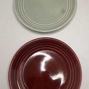 Peut inclure: Deux assiettes en céramique, une vert sauge et une bordeaux, sont présentées sur un fond blanc. Les deux assiettes sont circulaires avec des détails concentriques en relief. Ces assiettes sont adaptées pour servir des aliments.