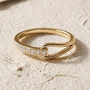 Può includere: Un anello d'oro dal design unico. L'anello presenta una fila di piccole pietre preziose a taglio quadrato incastonate in una sezione rettangolare. La fascia si curva e si collega alla sezione delle pietre preziose, creando un aspetto distintivo. L'anello è posto su una superficie strutturata bianco sporco.