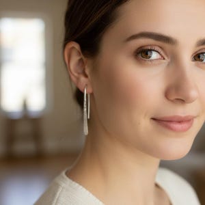May include: Silver-toned, long, dangling earrings. The earrings have a hammered texture and a linear design. The woman in the image is wearing a white sweater. The background is blurred.