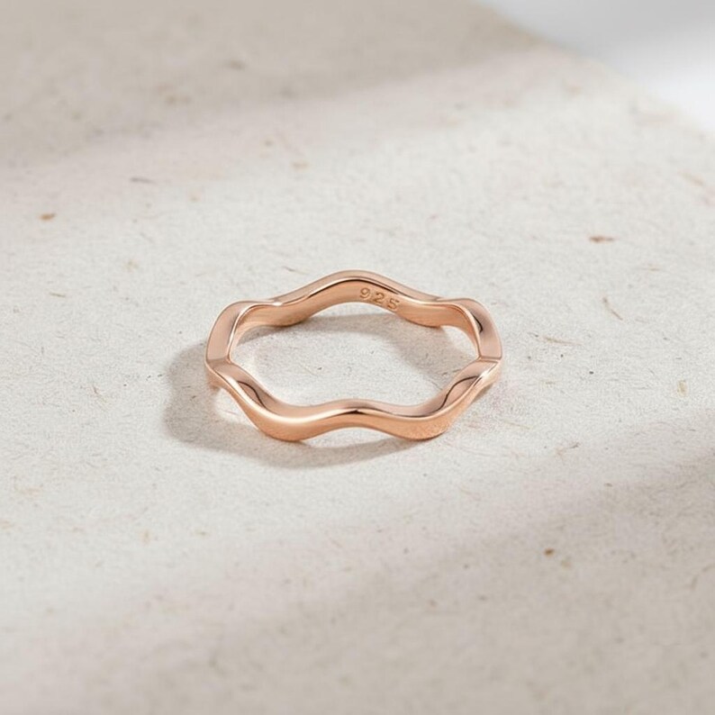 May include: A rose gold ring with a wavy design. The ring is made of metal and has a smooth, polished finish. The number "925" is stamped on the inside of the ring. The ring is a piece of jewelry.