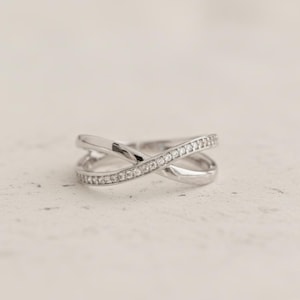 May include: A silver ring with an overlapping X design. One band is set with small, clear gemstones. The ring is displayed against a neutral background, highlighting its elegant and modern style. A piece of jewellery.