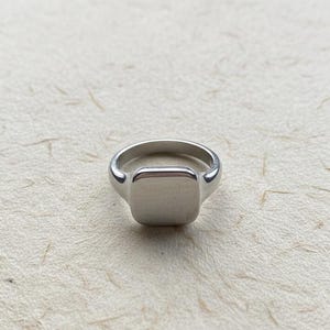 May include: A silver-colored signet ring with a square face. The ring has a smooth, polished finish and a rounded band. The ring is shown on a textured, off-white surface. This ring is a classic piece of jewelry.