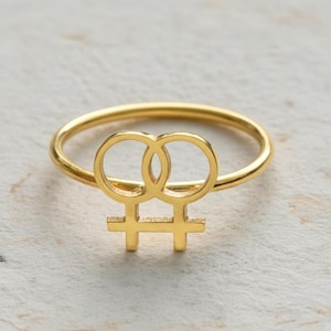 May include: A gold-colored ring featuring a stylized symbol of two interlocking circles above a horizontal line with two perpendicular lines. The ring is set against a light-colored background.