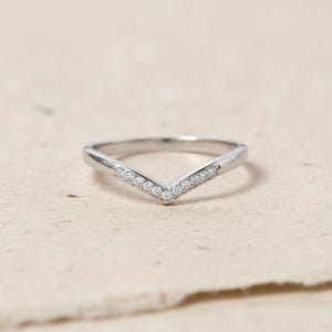 Pode incluir: Um anel prateado em forma de V com pequenas pedras preciosas transparentes ao longo do V. O anel está sobre uma superfície de tons neutros. O design do anel é simples e elegante, adequado para uso diário ou ocasiões especiais.
