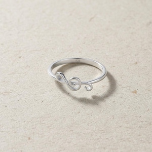May include: A silver ring featuring a treble clef design. The ring is made of a shiny, reflective metal and has a delicate, minimalist style. The ring is a music-themed accessory, perfect for musicians or music lovers.