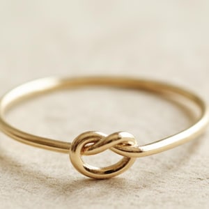 Puede incluir: Un delicado anillo dorado con un diseño de nudo en el centro. El anillo tiene una banda fina y un acabado pulido, creando una estética simple pero elegante. El detalle del nudo añade un toque único a la joya.