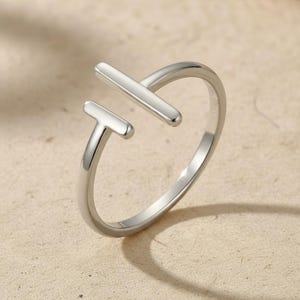 May include: A silver-colored ring with a modern, open design. The ring features two straight, parallel bars, one horizontal and one vertical, forming a T-shape. The ring is set against a neutral background, highlighting its minimalist aesthetic.