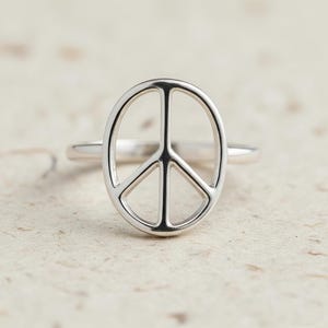 Könnte beinhalten: Ein silberfarbener Ring mit einem Peace-Zeichen. Der Ring hat eine polierte Oberfläche und ein schlichtes Band. Das Peace-Zeichen hat eine ovale Form mit einer vertikalen Linie und drei diagonalen Linien im Inneren. Der Ring wird vor einem neutralen Hintergrund gezeigt.