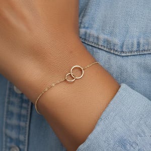 Könnte beinhalten: Ein zartes goldfarbenes Armband mit zwei ineinandergreifenden Kreisen. Das Armband ist an einer feinen Kette befestigt. Der Schmuck wird am Handgelenk getragen. Das Design ist schlicht und elegant, für den Alltag geeignet.