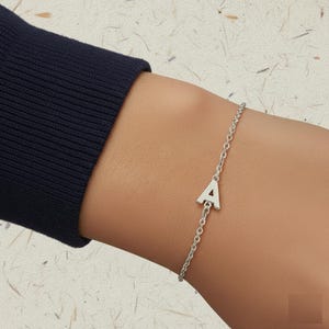 Op de afbeelding: Een zilveren kettingarmband met een letter "A" bedel. De armband is delicaat en eenvoudig, met een kleine, hoofdletter "A" hanger. De armband wordt om een pols gedragen, de achtergrond is licht en getextureerd.