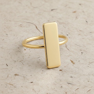 May include: A gold ring featuring a vertical rectangular bar. The ring has a polished finish and a minimalist design. The background is a textured, neutral tone. The ring is a simple, elegant piece of jewellery.