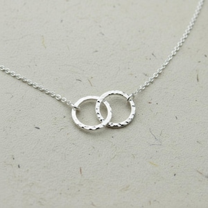 Può includere: Una delicata collana d'argento con due anelli martellati interconnessi. Gli anelli sono collegati da una sottile catena, creando un design minimalista ed elegante. La collana è esposta su una superficie strutturata di colore chiaro.