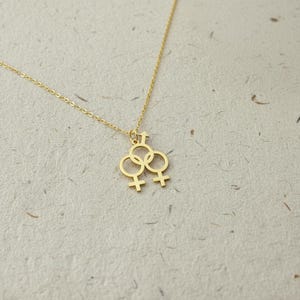 Könnte beinhalten: Eine goldfarbene Halskette mit einem Anhänger, der drei ineinandergreifende Geschlechtssymbole zeigt. Die Symbole umfassen eine Kombination aus männlichen und weiblichen Symbolen. Die Kette ist zart und der Anhänger ist zentriert.