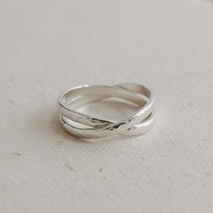 Puede incluir: Un anillo de plata con textura martillada. El anillo presenta un diseño entrecruzado, con dos bandas de metal que se superponen para crear una forma de X. El anillo está sobre un fondo neutro.