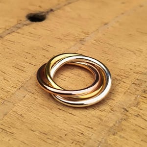 Op de afbeelding: Een driekleurige ring met in elkaar grijpende banden van goud, roségoud en zilver. De ring ligt op een houten oppervlak, waarbij de gepolijste afwerking en het contrast tussen de metalen worden benadrukt.