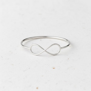 May include: A silver-colored ring featuring an infinity symbol design. The ring is made of a thin band with the infinity symbol at the center. The ring is a simple, elegant piece of jewelry.