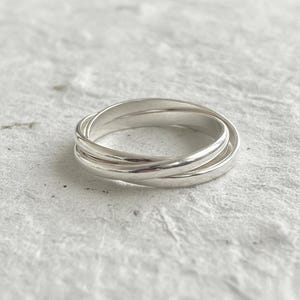 May include: A silver ring featuring three interlocking bands. The bands have a smooth, polished finish, reflecting light. The ring is displayed on a textured, off-white surface, emphasizing its simple design. A piece of jewellery.