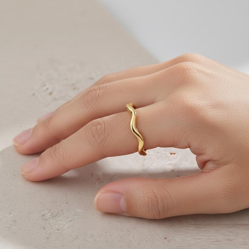 May include: A gold-colored ring with a wavy design. The ring is worn on a finger, showcasing its fluid, organic shape. The ring's surface reflects light, highlighting its polished finish. This piece of jewelry is a minimalist accessory.