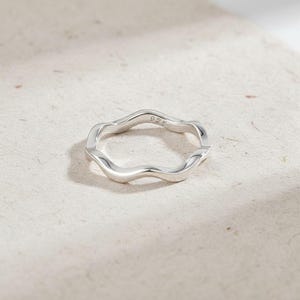 May include: A sterling silver ring with a wavy, organic design. The ring has a smooth, polished finish and features the marking "925" on the inside. The ring is displayed on a neutral-toned surface, highlighting its minimalist aesthetic.