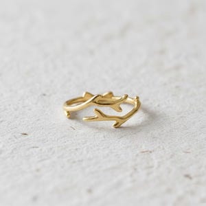 May include: A gold ring with a branch design. The ring has two curved branches that wrap around the finger. The ring is set against a white, textured background.