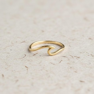 May include: A gold-colored ring in the shape of a wave. The ring has a smooth, polished finish and a minimalist design. The wave crest is curved and open, creating a delicate and elegant look. The ring is likely made of metal.