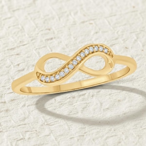 May include: A gold-colored ring in the shape of an infinity symbol. The infinity symbol is encrusted with small, clear gemstones. The ring is set against a textured, off-white background.