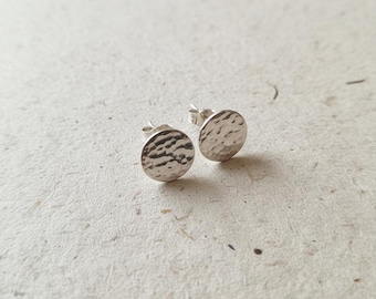 May include: A pair of silver stud earrings. Each earring is a small, hammered disc with a textured surface. The earrings are round and have a post for insertion into the ear. The earrings are displayed on a textured, off-white surface.