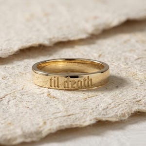 Peut inclure: Une bague anneau en or poli avec le texte gravé "til death" dans une police de style gothique. La bague présente une surface lisse et réfléchissante ainsi qu'une largeur constante, mettant en valeur les lettres détaillées gravées.