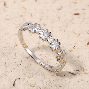 May include: A silver ring featuring a row of daisy flower designs. Each flower has a small, clear stone in the center. The ring is set against a neutral background, highlighting the bright metal and floral pattern.